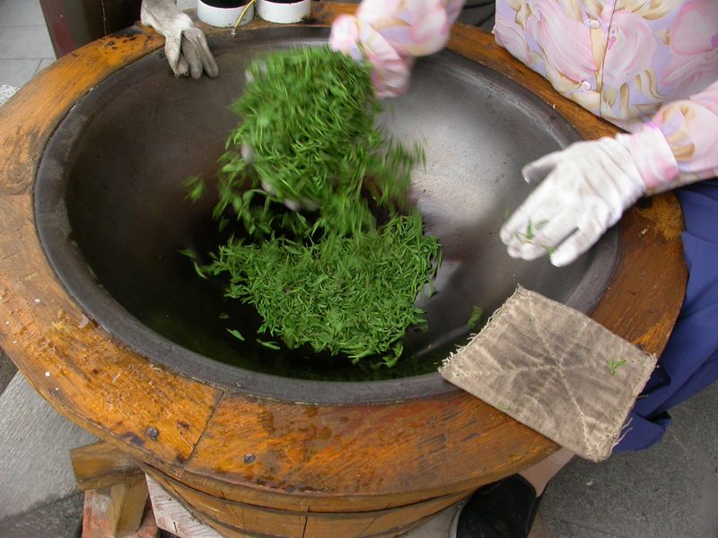 A woman in a conical hat kneeling at a Longjing Village tea house serving fresh Longjing in small glass cups