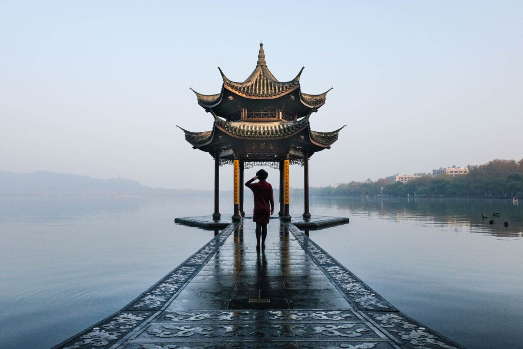 Solo female traveler in red dress standing on a stone path leading to a traditional Chinese pavilion on a misty Hangzhou lake